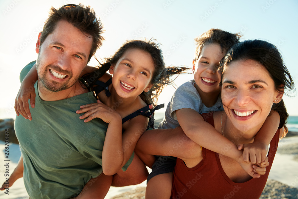 Mid adult white parents piggybacking their kids on a beach, smiling to camera, close up, backlit - obrazy, fototapety, plakaty 