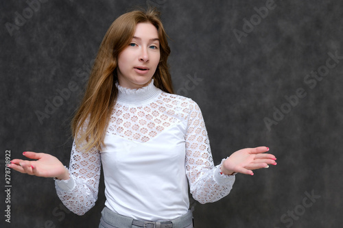 Portrait to the waist of a young pretty brunette girl woman with beautiful long hair on a gray background in a white jacket. He talks, smiles, shows his hands with emotions in various poses.