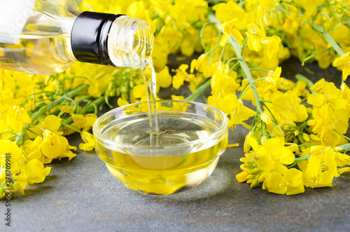 Pouring canola oil into the glass bowl against rapeseed blossoms on the grey surface, closeup shot photo