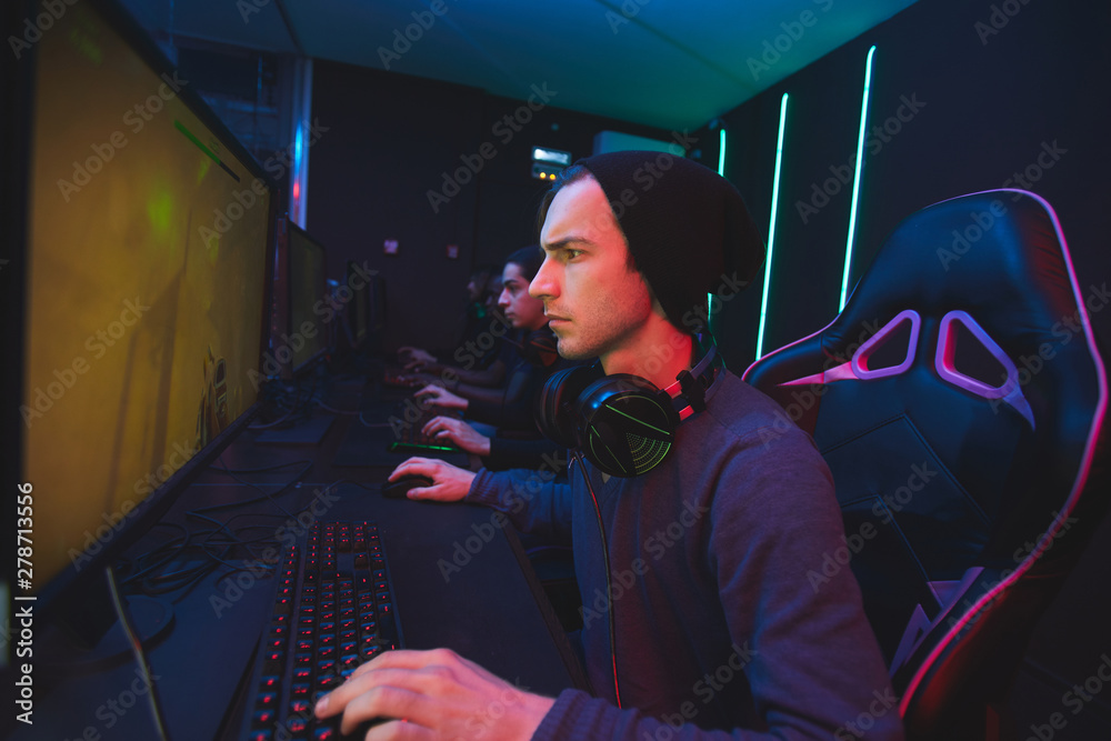 Fototapeta premium Serious young man in beanie sitting at table and using computer while playing shooter video game