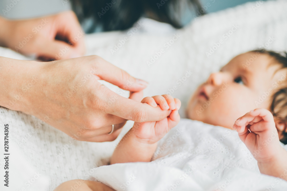 Adorable two month old baby girl lying on the bad. Mother holding daughter hand and smiling. Mother standing near the cradle and holding baby's hand. Concept photo parenthood and motherhood. - obrazy, fototapety, plakaty 