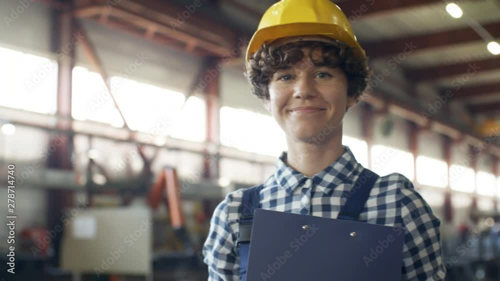 Pan chest-up shot of curly lady factory technician talking on walkie-talkie then looking at camera with relaxed and confident smile, with blurred spacious sunlit workshop in background Pan chest-up shot of curly lady factory technician talking on walkie-talkie then looking at camera with relaxed and confident smile, with blurred spacious sunlit workshop in background