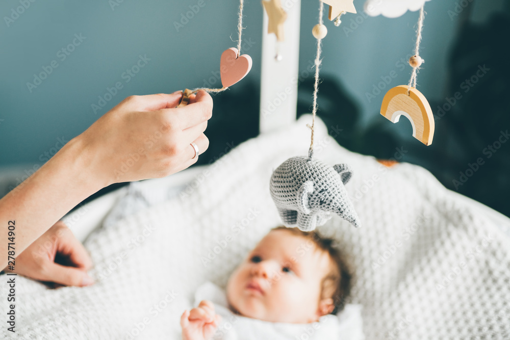 Adorable two month old baby girl lying on the bad. Mother standing near the cradle and holding clack. Concept photo parenthood and motherhood. - obrazy, fototapety, plakaty 