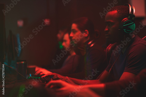 Group of concentrated multi-ethnic esport players sitting in front of computers in dark room full of red light and playing online games