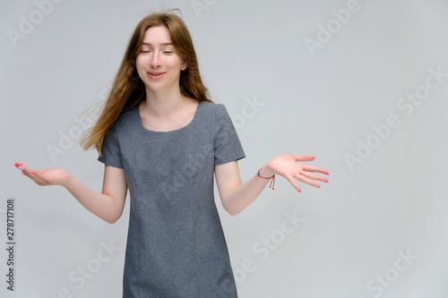 A portrait below belt of a young pretty brunette girl woman with beautiful long hair on a white background in a gray dress. He talks, smiles, shows his hands with emotions in various poses.