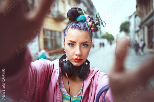 Playful cool funky hipster young girl with headphones and crazy hair taking selfie on street photo