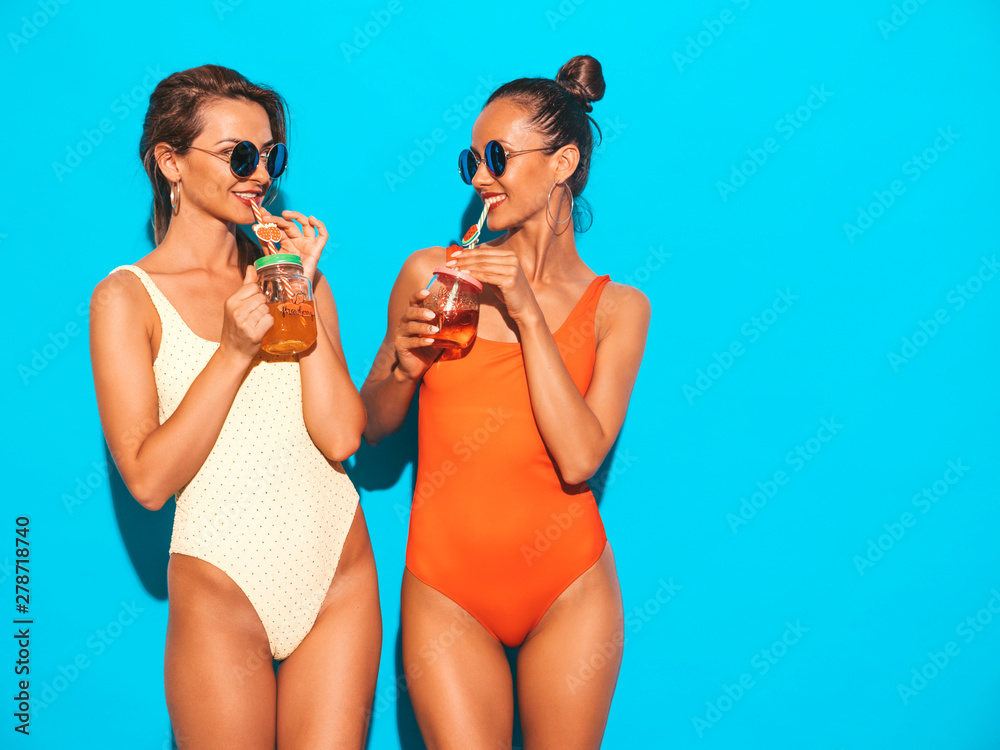 Two beautiful sexy smiling hipster women in summer colorful swimwear bathing suits.Trendy girls in sunglasses.Going crazy.Funny models isolated on blue.Drinking fresh cocktail smoozy drink Two beautiful sexy smiling hipster women in summer colorful swimwear bathing suits.Trendy girls in sunglasses.Going crazy.Funny models isolated on blue.Drinking fresh cocktail smoozy drink