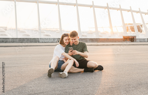 Beautiful couple sitting on the street, looking at the smartphone screen and smiling, backdrop of the sunset and modern architecture.Happy couple in love are watching funny videos on the smartphone