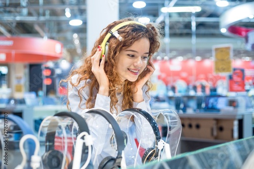 Red-haired girl standing in front of the stand in the electronics store chooses headphones before buying wearing on her head listening to the sound