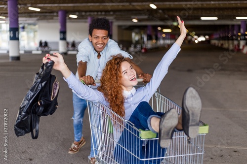 African guy wheels happy redheaded girl in the grocery cart on sale in the supermarket Parking in the centre targowa photo