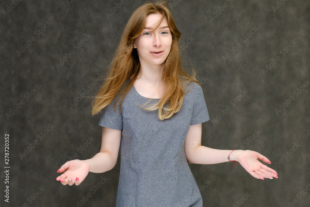 Portrait to the waist of a young pretty brunette girl woman with beautiful long hair on a gray background in a gray dress. He talks, smiles, shows his hands with emotions in various poses. Portrait to the waist of a young pretty brunette girl woman with beautiful long hair on a gray background in a gray dress. He talks, smiles, shows his hands with emotions in various poses.