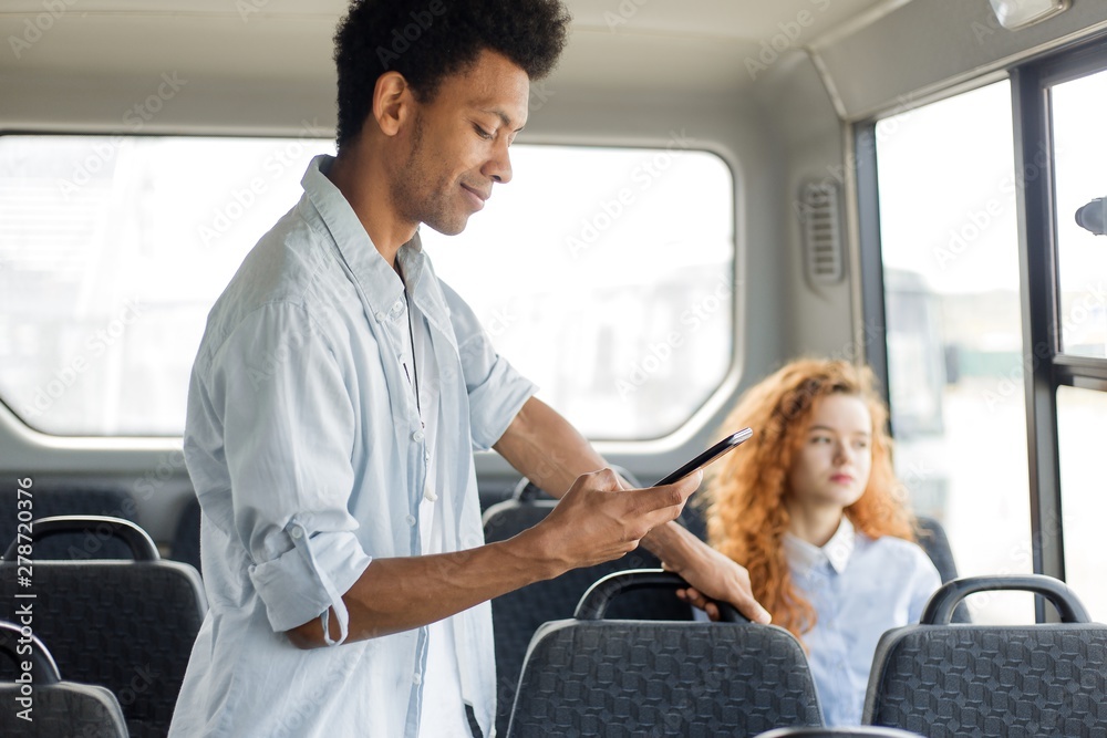 Passengers traveling in public transport. African man with a phone in his hands holding the handrail standing next to a sitting red-haired girl riding the bus. View inside the bus Passengers traveling in public transport. African man with a phone in his hands holding the handrail standing next to a sitting red-haired girl riding the bus. View inside the bus