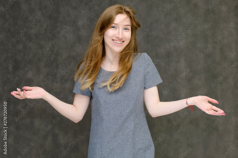 Portrait to the waist of a young pretty brunette girl woman with beautiful long hair on a gray background in a gray dress. He talks, smiles, shows his hands with emotions in various poses. Portrait to the waist of a young pretty brunette girl woman with beautiful long hair on a gray background in a gray dress. He talks, smiles, shows his hands with emotions in various poses.