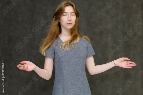 Portrait to the waist of a young pretty brunette girl woman with beautiful long hair on a gray background in a gray dress. He talks, smiles, shows his hands with emotions in various poses.