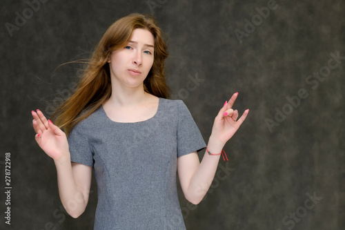 Portrait to the waist of a young pretty brunette girl woman with beautiful long hair on a gray background in a gray dress. He talks, smiles, shows his hands with emotions in various poses.