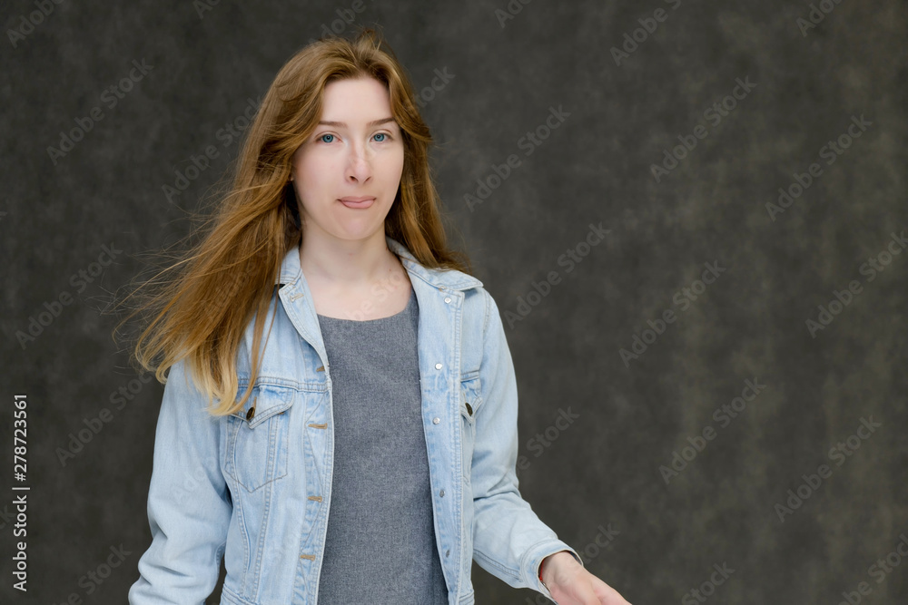 Waist up portrait of a young pretty brunette girl woman with beautiful long hair on a gray background in a jeans jacket. He talks, smiles, shows his hands with emotions in various poses. Waist up portrait of a young pretty brunette girl woman with beautiful long hair on a gray background in a jeans jacket. He talks, smiles, shows his hands with emotions in various poses.