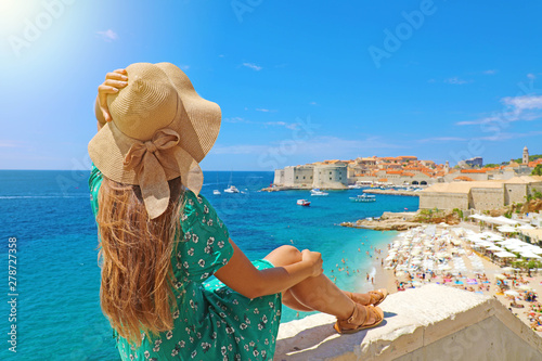 Beautiful young woman sitting on wall looking at stunning view of Mediterranean sea and Dubrovnik old town in Croatia, Europe. Lifestyle woman with straw hat wearing green dress enjoy landscape view.