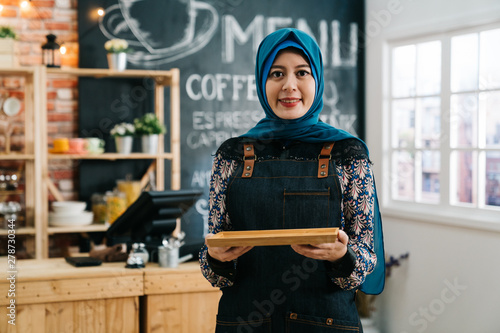 Female Muslim Cafe Worker Greeting Customer In Coffee Shop Young Malay Girl In Apron Holding Tray