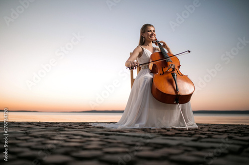 Girl in a white dress playing the cello on the shore of the lake, after sunset. There is a place for text, perfect for a cover or poster, advertising or signage.