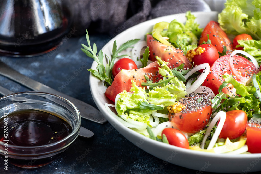 Salad of tomato, onion, pepper, arugula and chia seeds. Dressed with soy sauce and olive oil. Near the stick with dressing. On a dark background. Metal fork and knife. Salad of tomato, onion, pepper, arugula and chia seeds. Dressed with soy sauce and olive oil. Near the stick with dressing. On a dark background. Metal fork and knife.