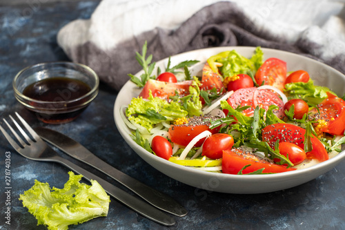 Salad of tomato, onion, pepper, arugula and chia seeds. Dressed with soy sauce and olive oil. Near the stick with dressing. On a dark background. Metal fork and knife. 