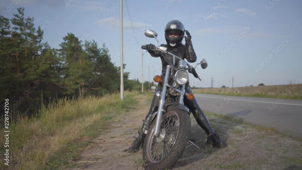 Front view of the confident young woman in leather dress sitting on her bike at the road. Hobby, traveling and active lifestyle. Female biker outdoors on her motorbike. Slow motion.