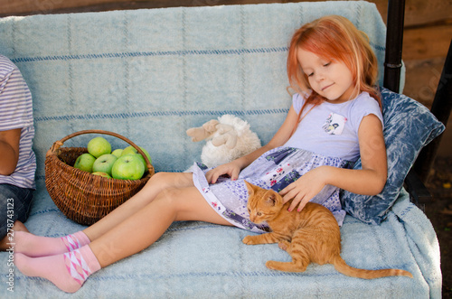 Ginger kitten lies near the girl on the garden swing. The girl strokes her pet. Little blonde caucasian girl with a red cat