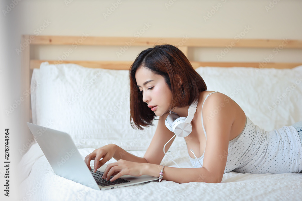 close up shot of beautiful and sexy young adult asian woman in white dress lying on bed in bedroom while using computer and listening music from headphone close up shot of beautiful and sexy young adult asian woman in white dress lying on bed in bedroom while using computer and listening music from headphone