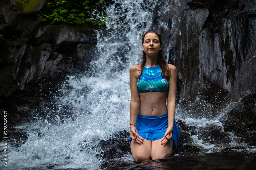 Young Caucasian woman meditating, practicing yoga at waterfall in Ubud, Bali, Indonesia.