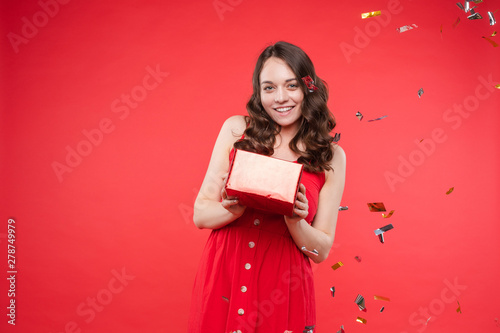 Front view of happy young girl wearing red dress looking at camera and twirling hair with hand in studio. Curly model posing and smiling on isolated background. Concept of emotions and celebration.