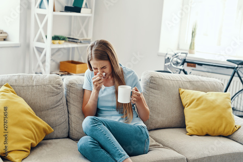 Canvas Print Ill young woman blowing nose and sneezing while sitting on the sofa at home