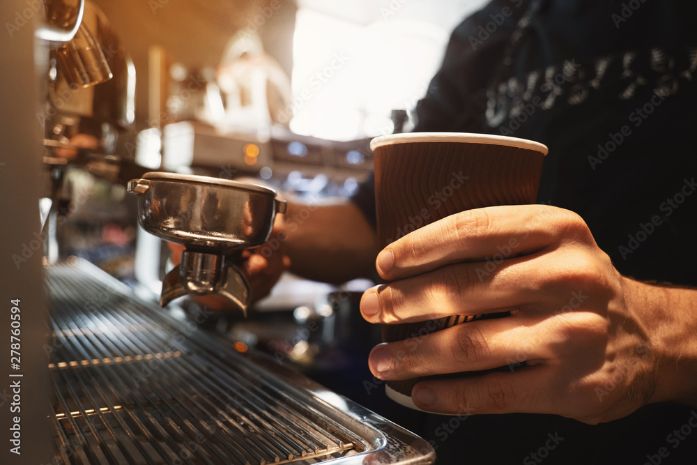 barista man holding hot coffee drink in paper cup in one hand and coffee holder in another standing close to professional coffee machine in cafe close up