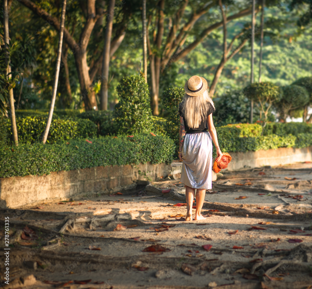 Blonde in a straw hat and a long skirt walks through a tropical park. Back view. Thailand travel Blonde in a straw hat and a long skirt walks through a tropical park. Back view. Thailand travel