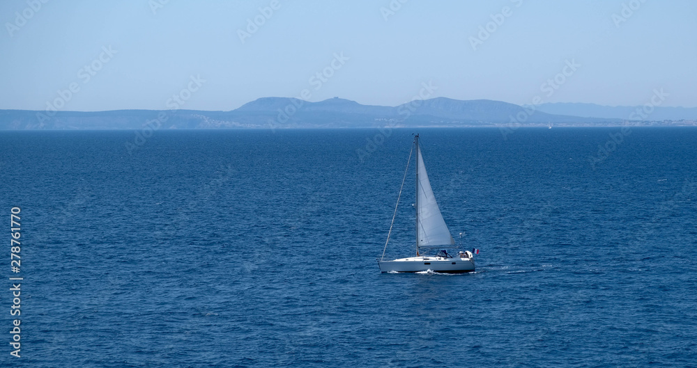 Small sailboat at the sea. Sailing near the shore in the mediterranean sea. Amazing sunny day, calm blue water. Sail race in the coastal bay. Boat in the sea. Happy vacation. White sail.