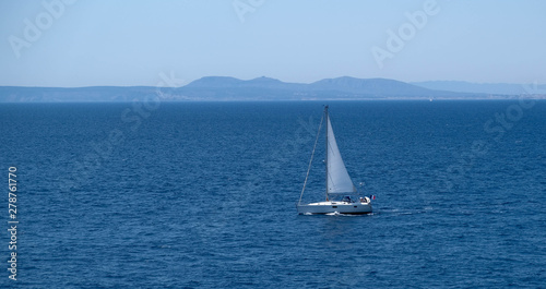 Small sailboat at the sea. Sailing near the shore in the mediterranean sea. Amazing sunny day, calm blue water. Sail race in the coastal bay. Boat in the sea. Happy vacation. White sail.