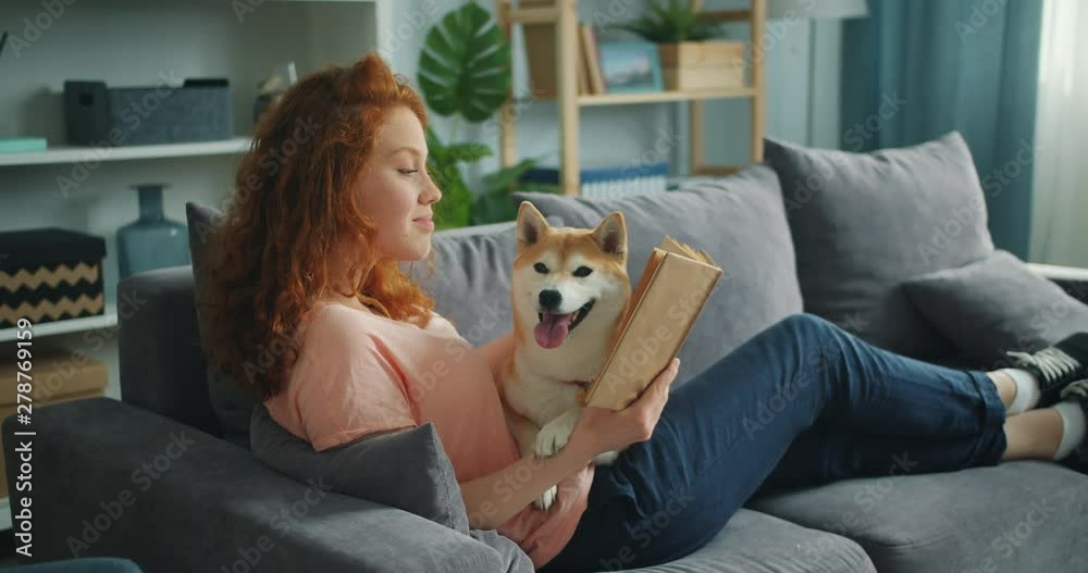 Pretty student young woman is reading book in cozy apartment smiling and petting adorable dog sitting on comfy couch at home. Animals and hobby concept.