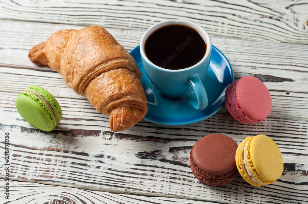 One whole baked croissant near various macaroons and full blue ceramic cup of fresh black coffee on old white rustic wooden table One whole baked croissant near various macaroons and full blue ceramic cup of fresh black coffee on old white rustic wooden table