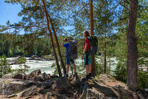Kids travel hiking in nature. Boys with backpacks look through binoculars at the mountain river.