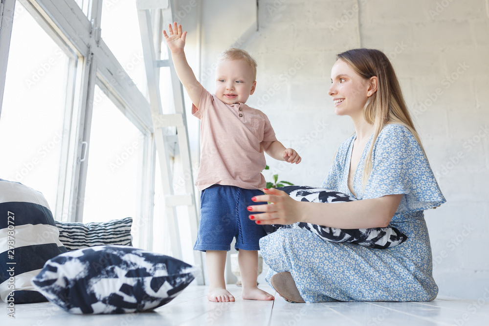 Φωτογραφία Side view of cheerful woman holding baby boy in her hands and looking at him with love sitting on wooden white floor over big window at home