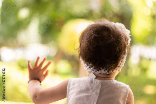 cute baby girl in a white gown leaning on a window glass and looking outside on a green park, while waiting for her parents to come back to home.