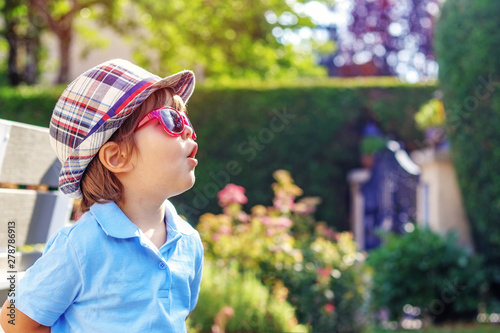 Portrait of funny curious little boy in hat and sunglasses looking surprised with open mouth outdoors in garden at sunny summer day. Summer lifestyle.