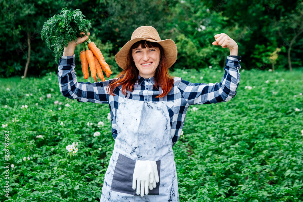 Portrait beautiful female farmer holds a bunch of carrots smiling at the camera wearing a straw hat and surrounded by the many plants in her vegetable garden