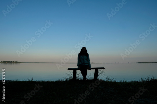 Silhouette of a meditating woman 59 years old at dawn on Svityaz Lake in Ukraine. The woman is sitting on a home-made wooden bench. Self-isolation concept.