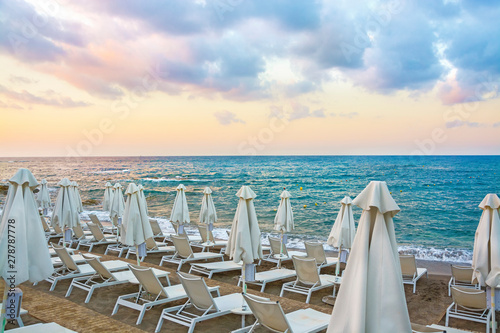 Sun beds and umbrellas on an empty beach in the background of a beautiful cloudy sky in Crete Island, Greece. Concept of summer holiday, vacation and tourism. © MarinadeArt