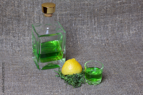 Strong tincture on tarhuna in a transparent rectangular bottle. Drink bright green poured into a glass. Next to the ingredients for cooking. Tarragon leaves and lemon. photo