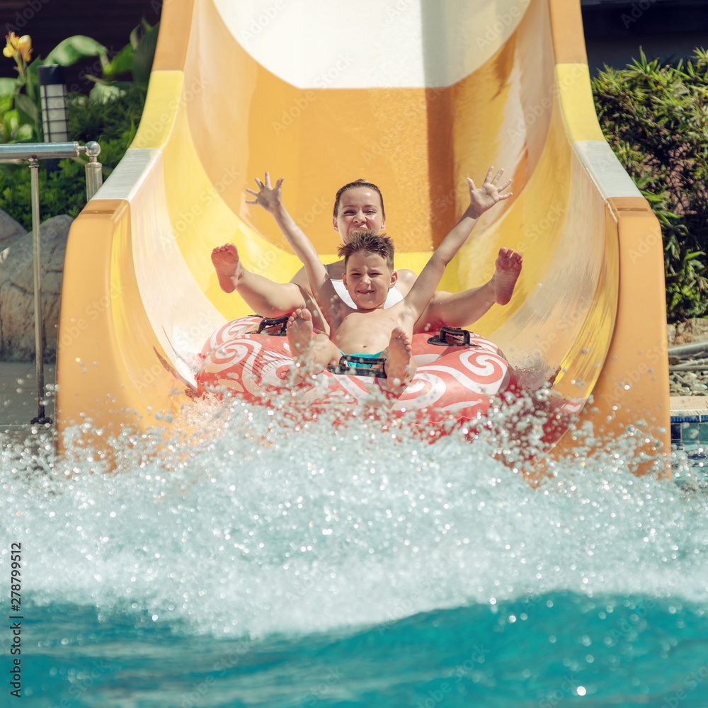 Caucasian boy and mom gliding down slide in waterpark. They enjoy the fun and holding hands wide open.