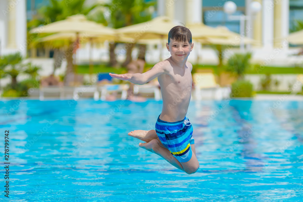 European boy in striped swimming shorts is having fun jumping into swimming pool at resort.