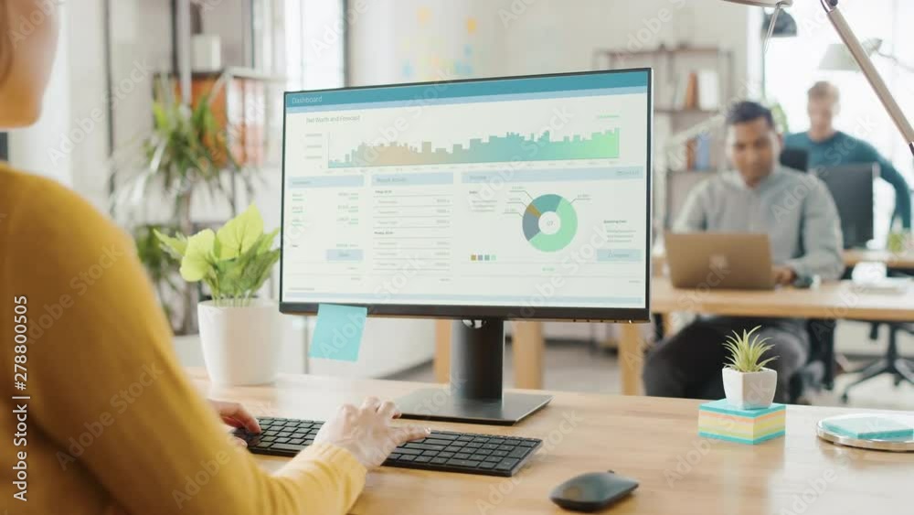 Over the Shoulder: Creative Young Woman Sitting at Her Desk Using Desktop Computer with Screen Showing Project Management, Statistics and Graphs. Office with Diverse Team of Professionals Working