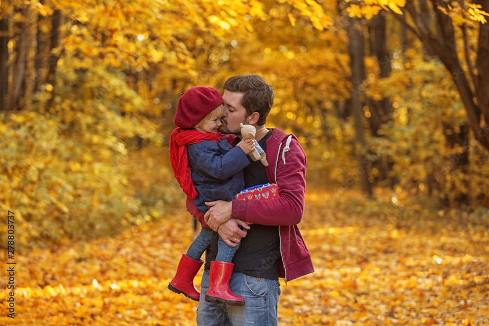 Father hugs and kisses his daughter in autumn day. The girl is holding a teddy bear.