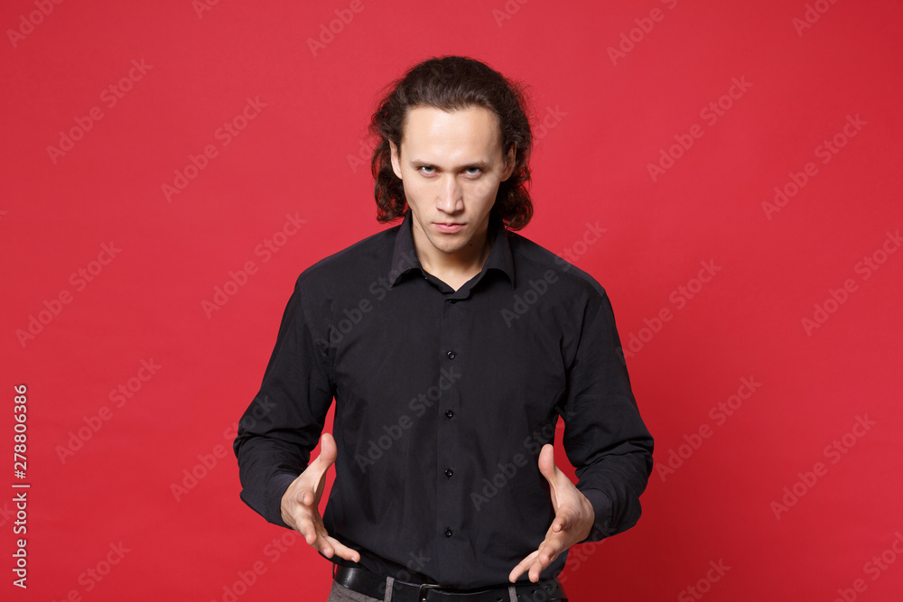 Fototapeta premium Shocked upset stylish young curly long haired man in black shirt posing isolated on red wall background studio portrait. People sincere emotions lifestyle concept. Mock up copy space. Looking camera.